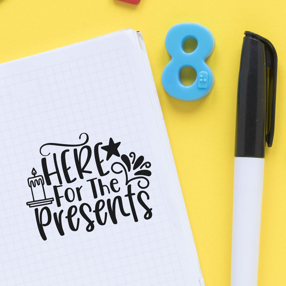 Blue number 8 stamp and black marker beside a notebook with a black Here for the presents seal on a yellow background.