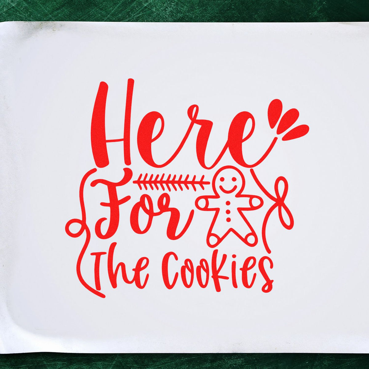 Red cookie stamp reading Here For The Cookies with gingerbread man on white sheet, bright red on green background