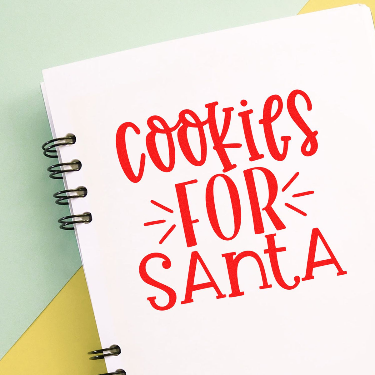 Red COOKIE FOR SANTA stamp on white spiral-bound notebook, with black rings on a pastel green and yellow background.