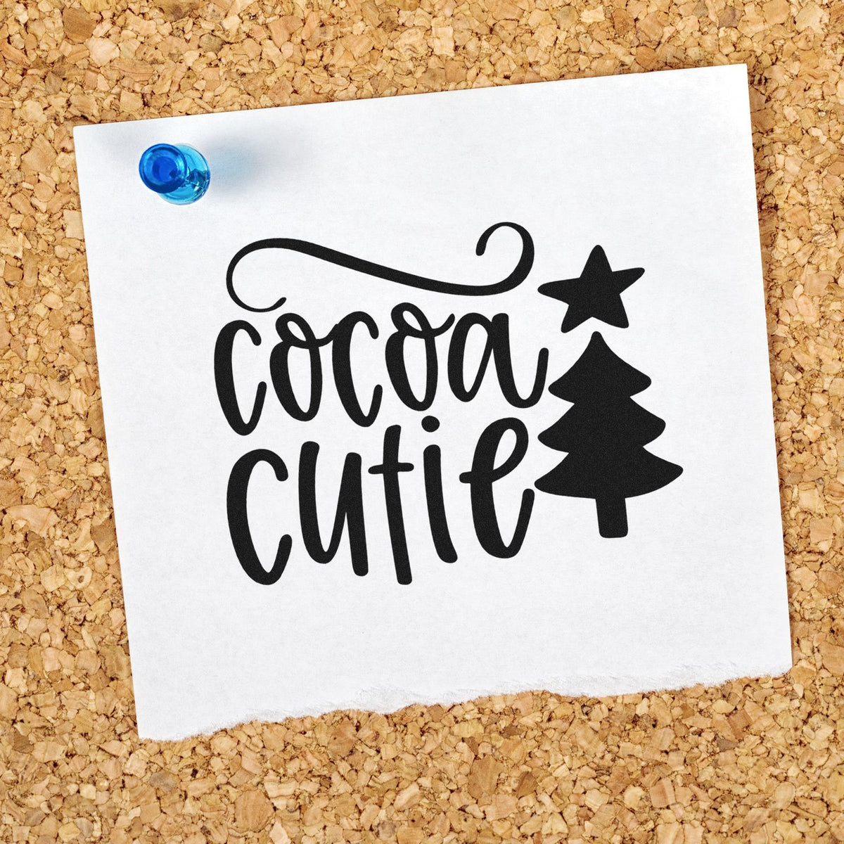 Black holiday stamp reading cocoa cutie with star and pine tree, pinned to corkboard with a blue pushpin.