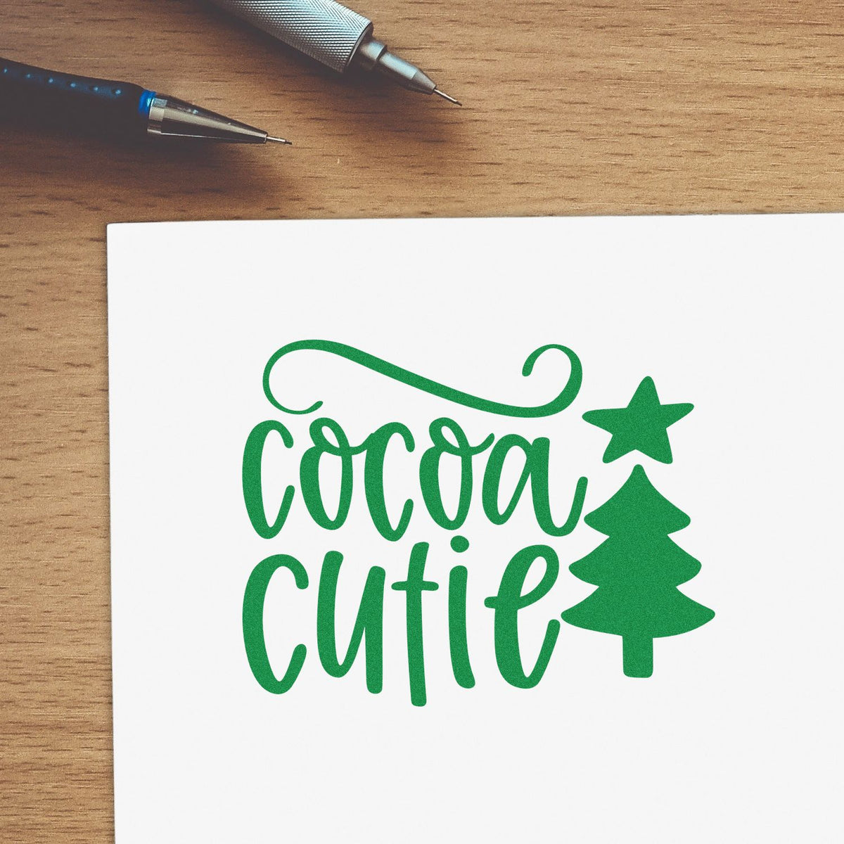 Green Cocoa Cutie custom stamp and seal design with Christmas tree and star on white paper, beside silver and black pens.