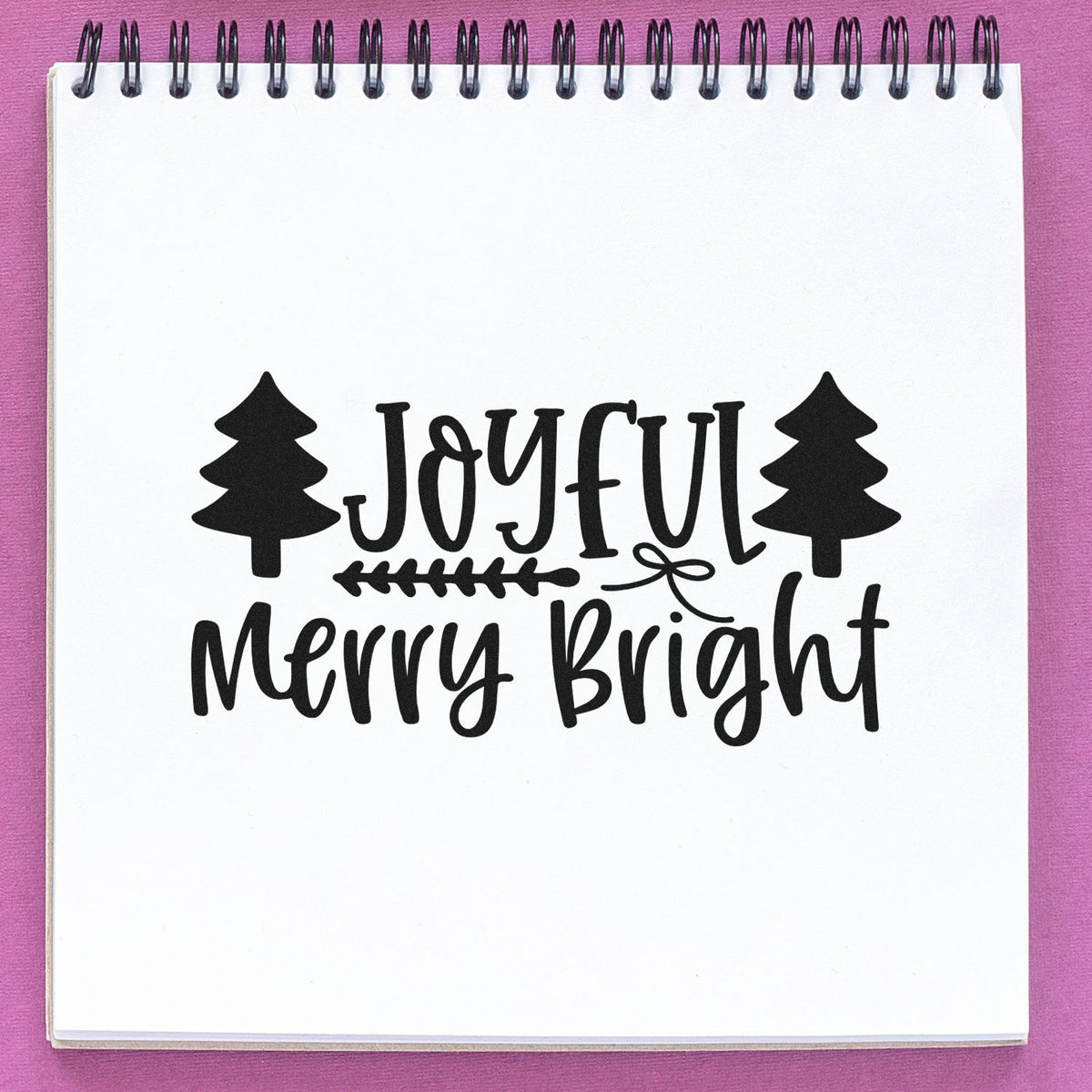 Black Joyful Merry Bright holiday stamp with pine trees on white spiral notebook paper, set against a pink background.