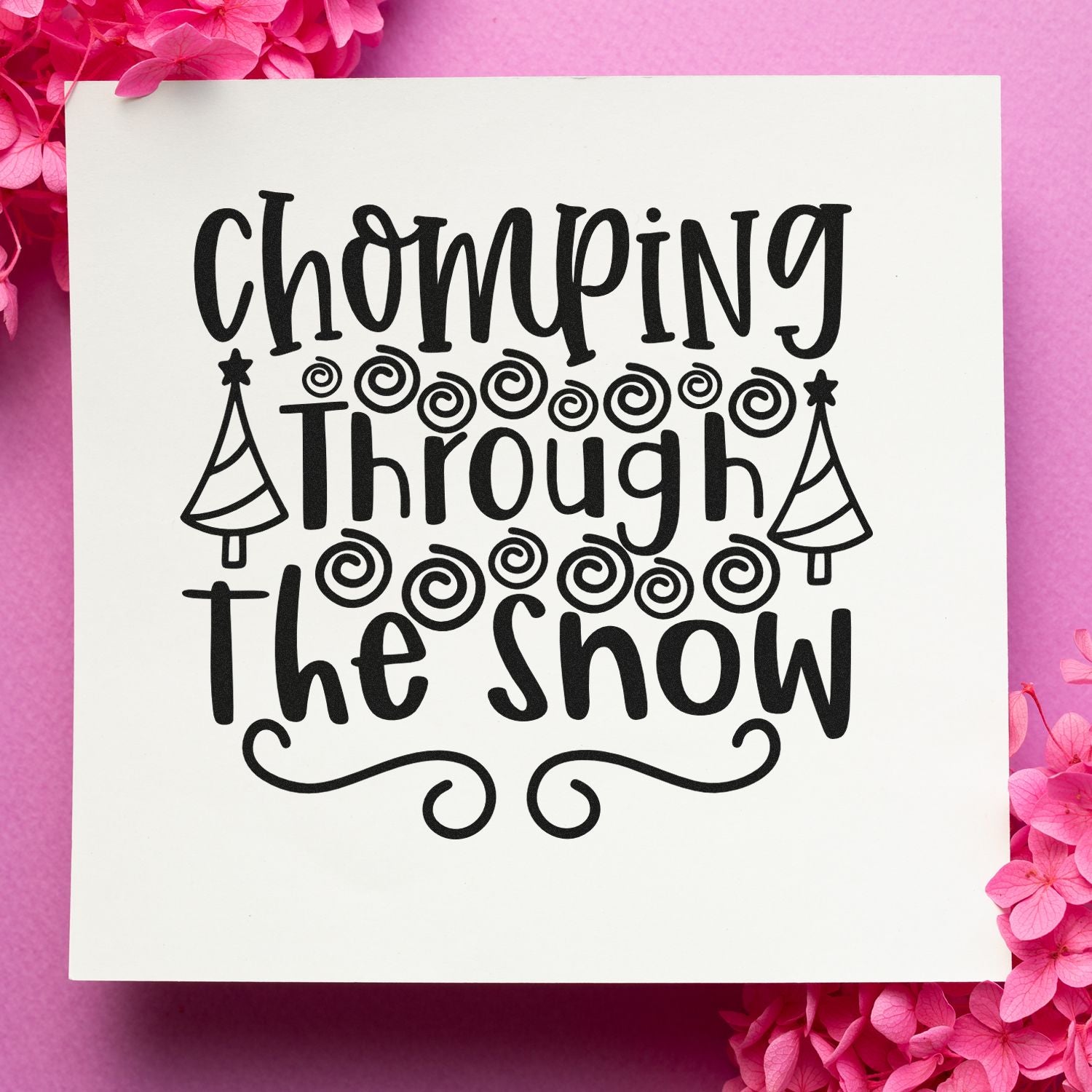 Black decorative stamp and seal reading "Chomping Through the Snow" with trees and swirls on a white background, framed by pink flowers.