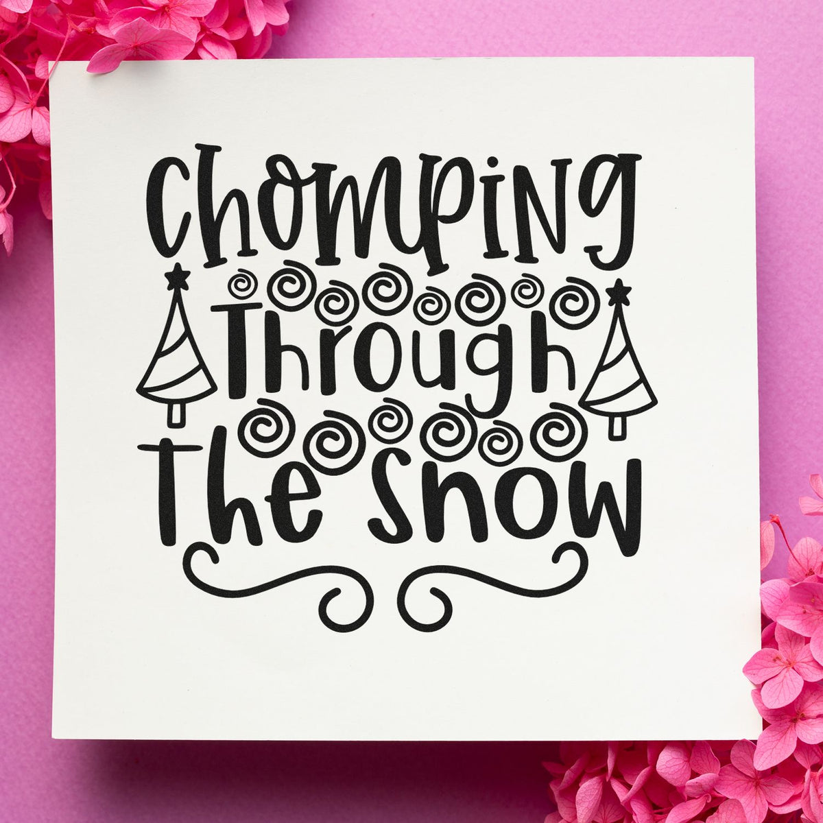 Black stamp and seal design reading Chomping Through the Snow with swirls, trees, on white background and pink flowers.