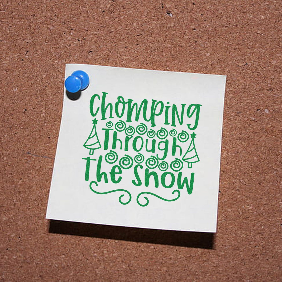 Green holiday stamp and seal design reading "Chomping Through the Snow" with trees and swirls on white paper pinned to corkboard.