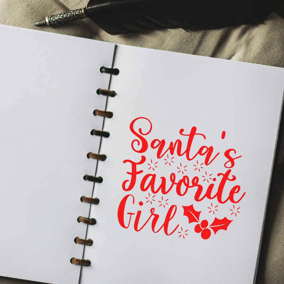 White notebook page with bright red Santa's Favorite Girl holiday stamp/seal, holly accents, spiral binding, and pen