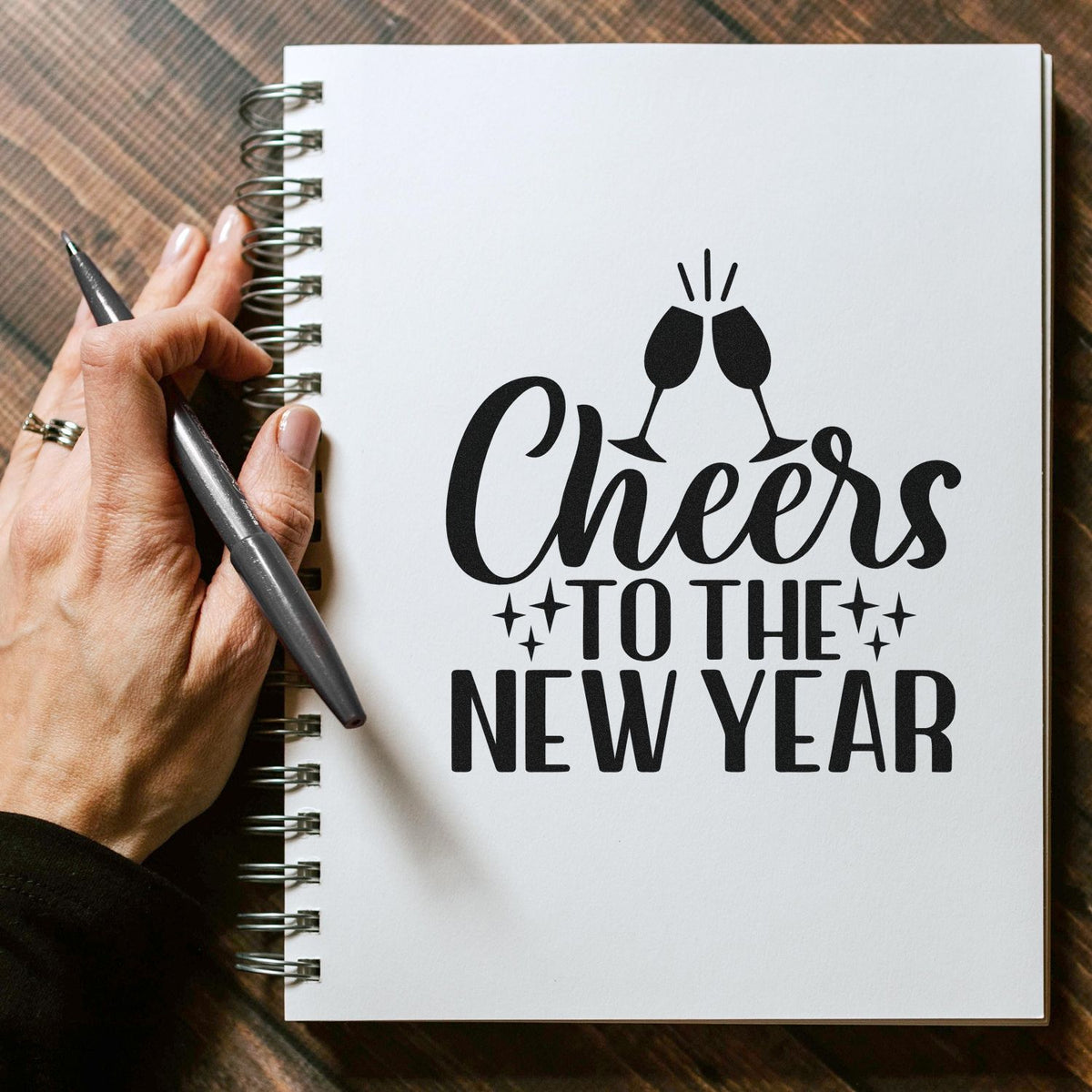 Black Cheers to the New Year stamp/seal with clinking champagne glasses on a white spiral notebook, held by a hand with pen.