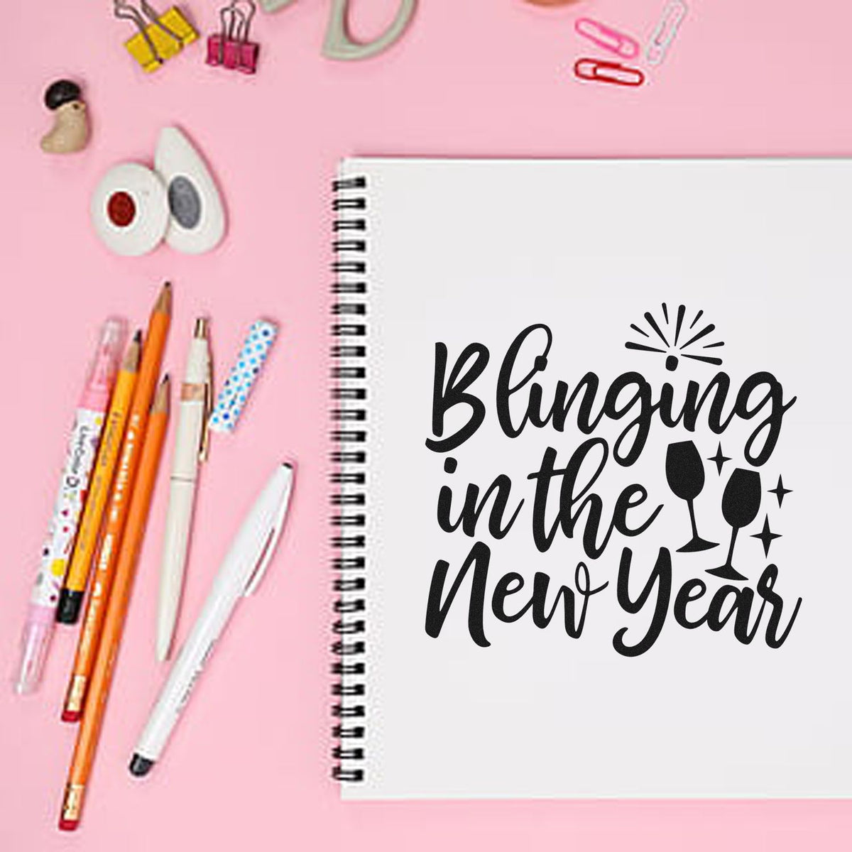 Pink flat lay of stamp and seal products, spiral notebook stamped Blinging into the New Year with black wine glass icons.
