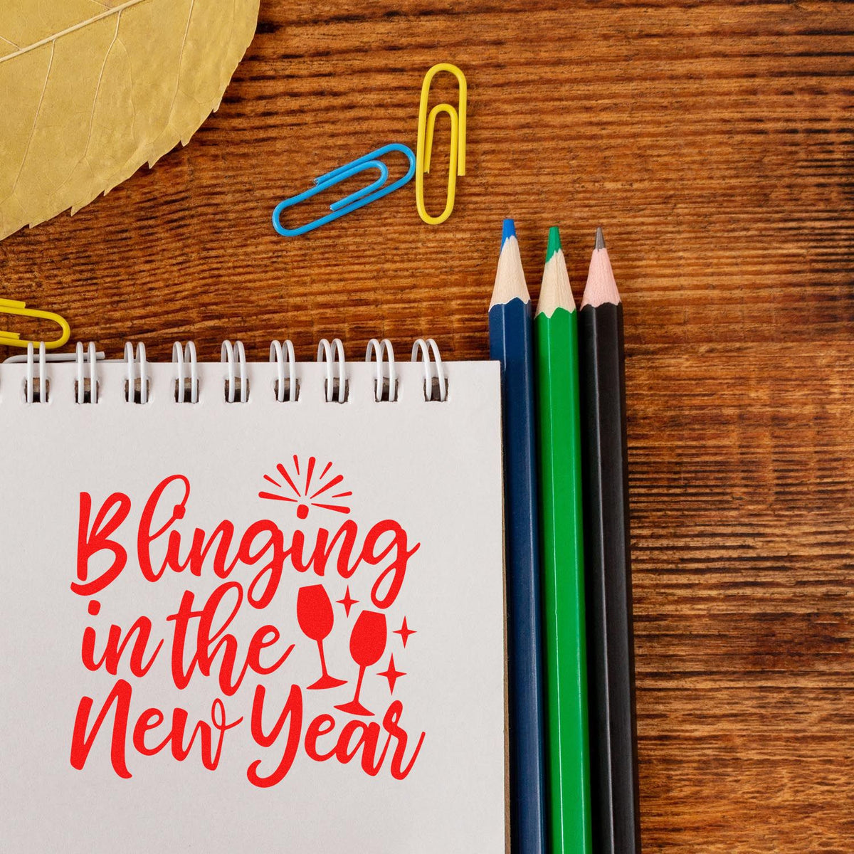 Red New Year stamp and seal design on a white notebook, with champagne flutes, colored pencils, and paper clips on wood.