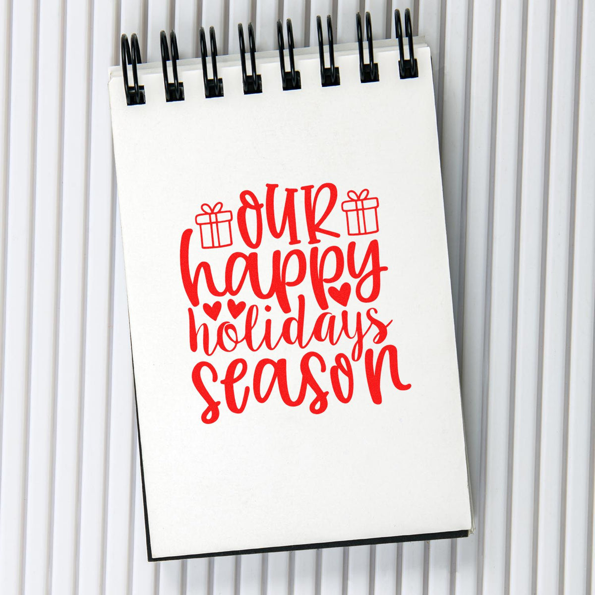 Red Our Happy Holidays Season stamp design on white spiral notepad with gift icons and hearts.