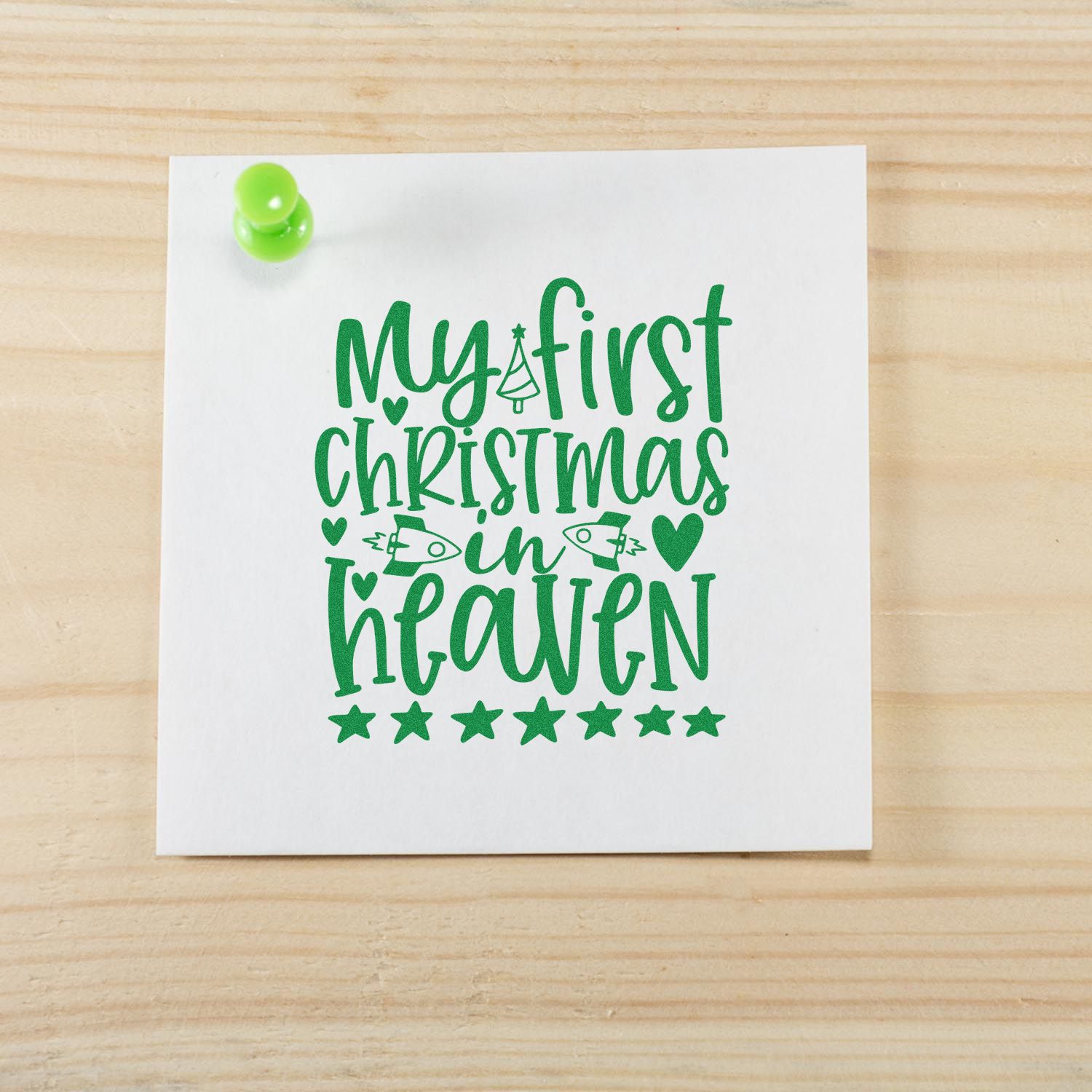 Green stamp design on white card with pushpin, reading My first Christmas in heaven with hearts, stars and trees.
