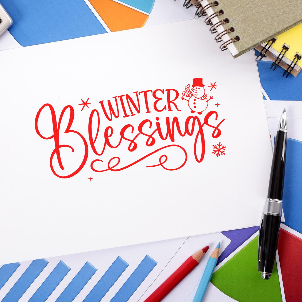 Red Winter Blessings stamp design with snowman on white paper, framed by colorful stationery and pens.