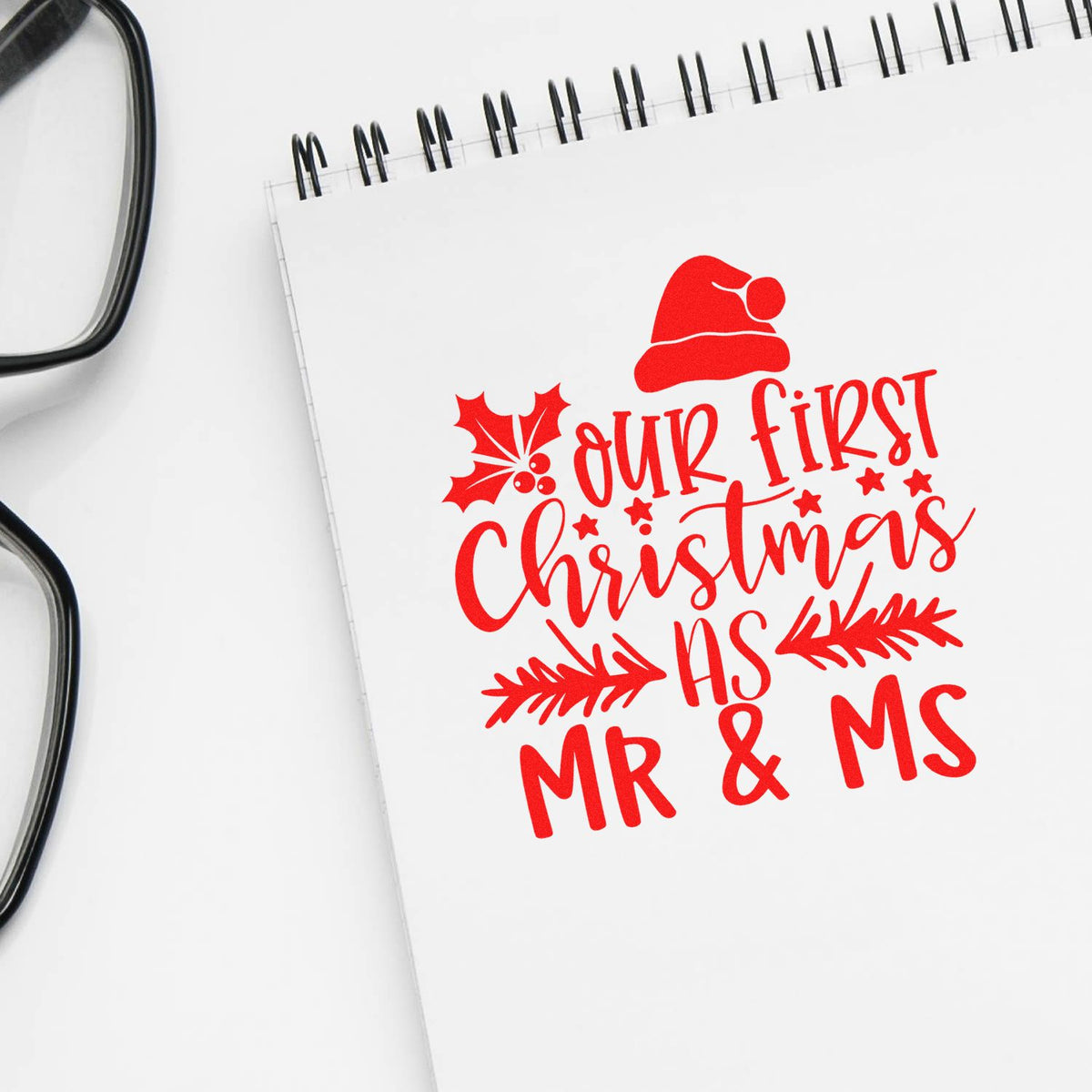 Red holiday stamp design on white notebook: Our First Christmas as Mr & Ms with Santa hat, holly, and stars.