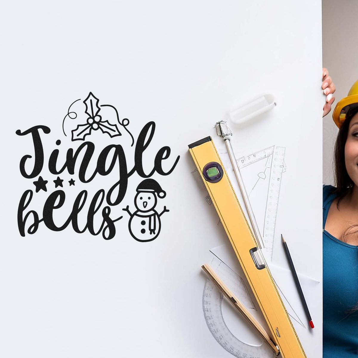 Black Jingle Bells holiday seal stamp with holly and snowman on white board, beside yellow level and drafting tools.