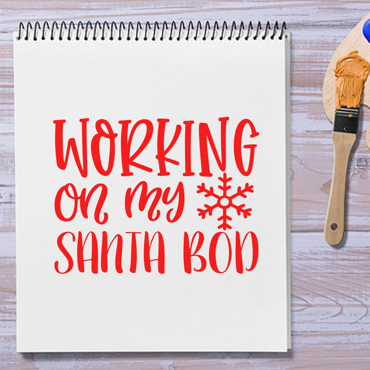 Red holiday stamp and seal on white notebook reading Working on my Santa bod, with snowflake and wooden brush.