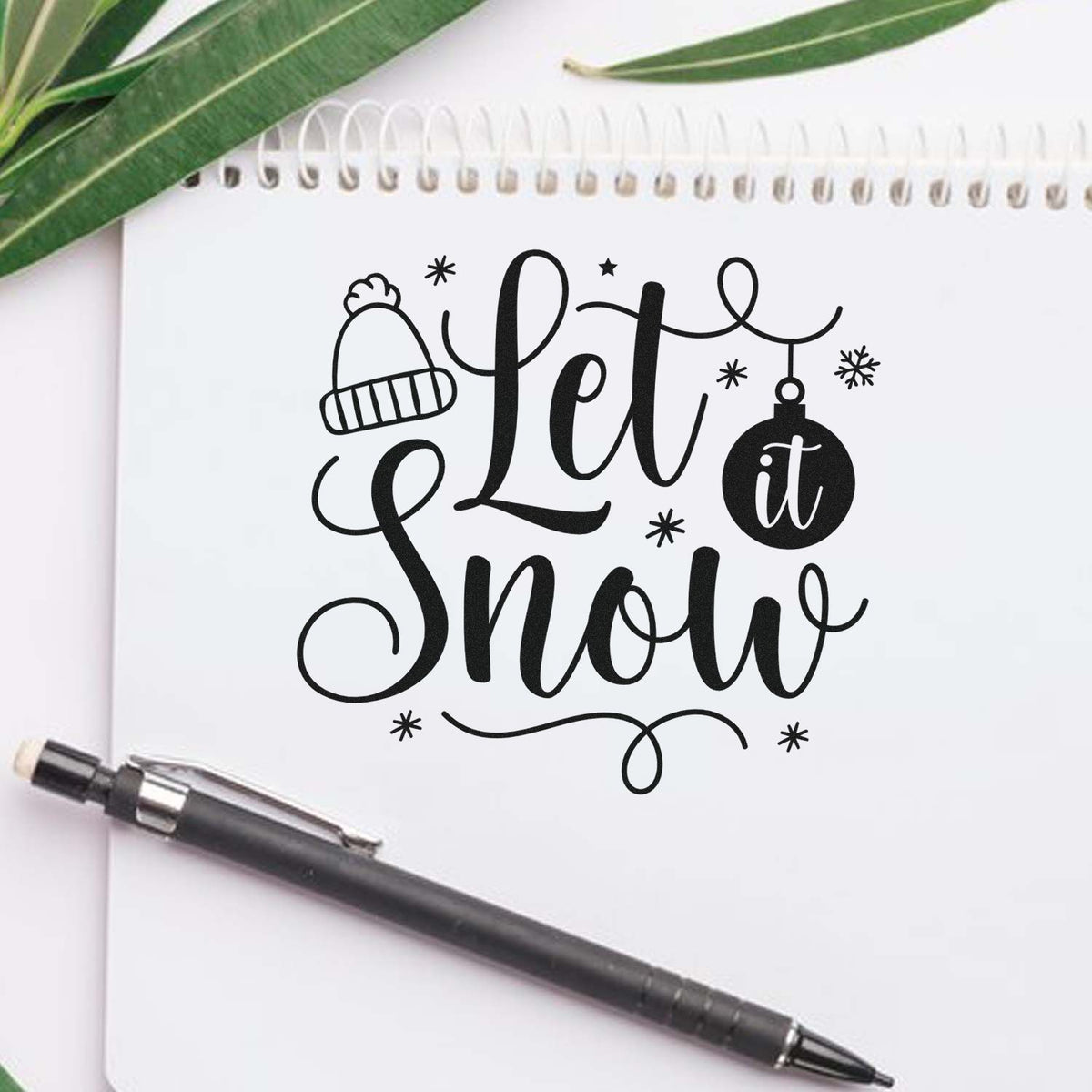 Black winter stamp and seal reading Let it Snow with beanie, ornament and snowflakes on a white notebook beside a pen.
