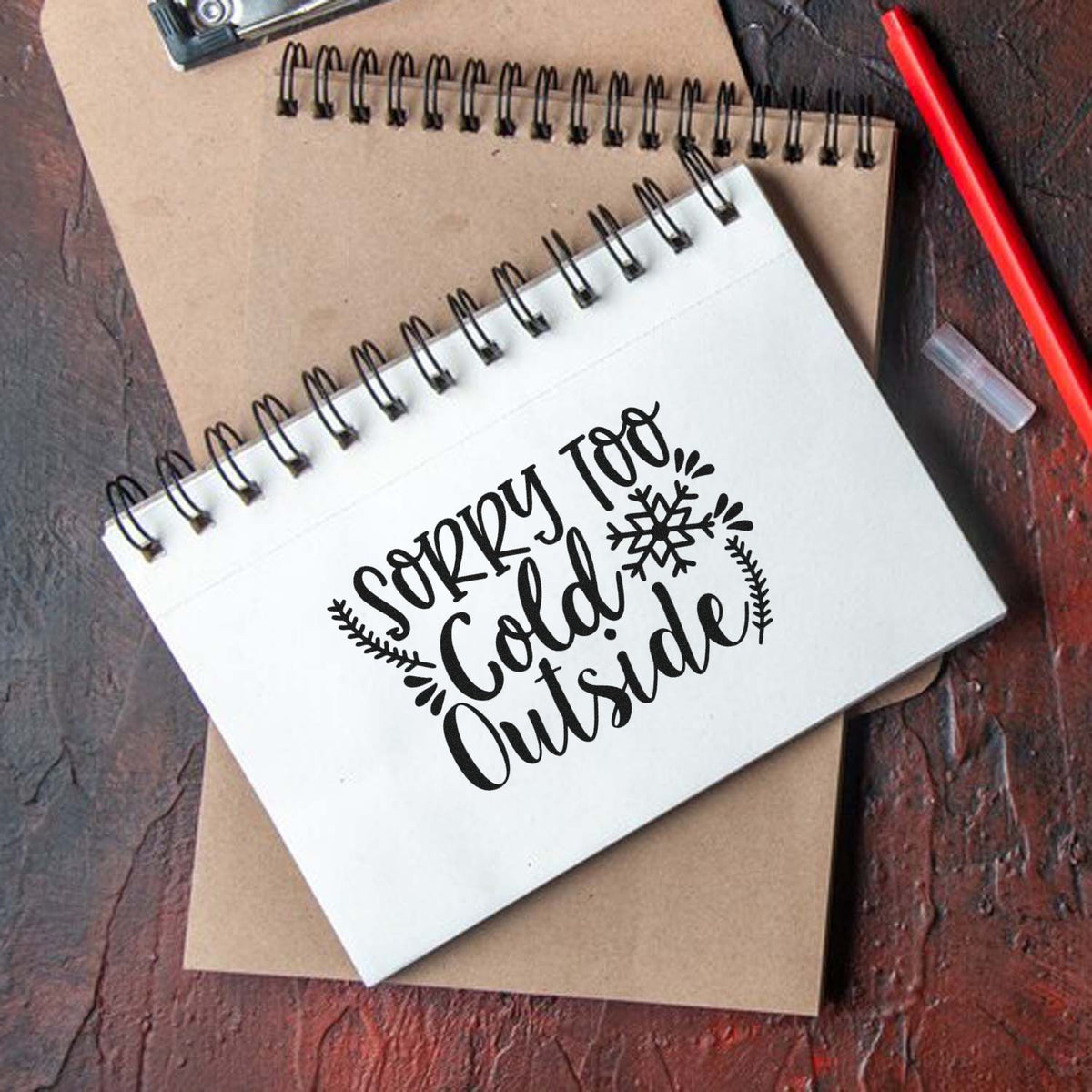 Black custom stamp/seal reading Sorry Too Cold Outside on a white spiral notebook, with snowflake accents and kraft paper.