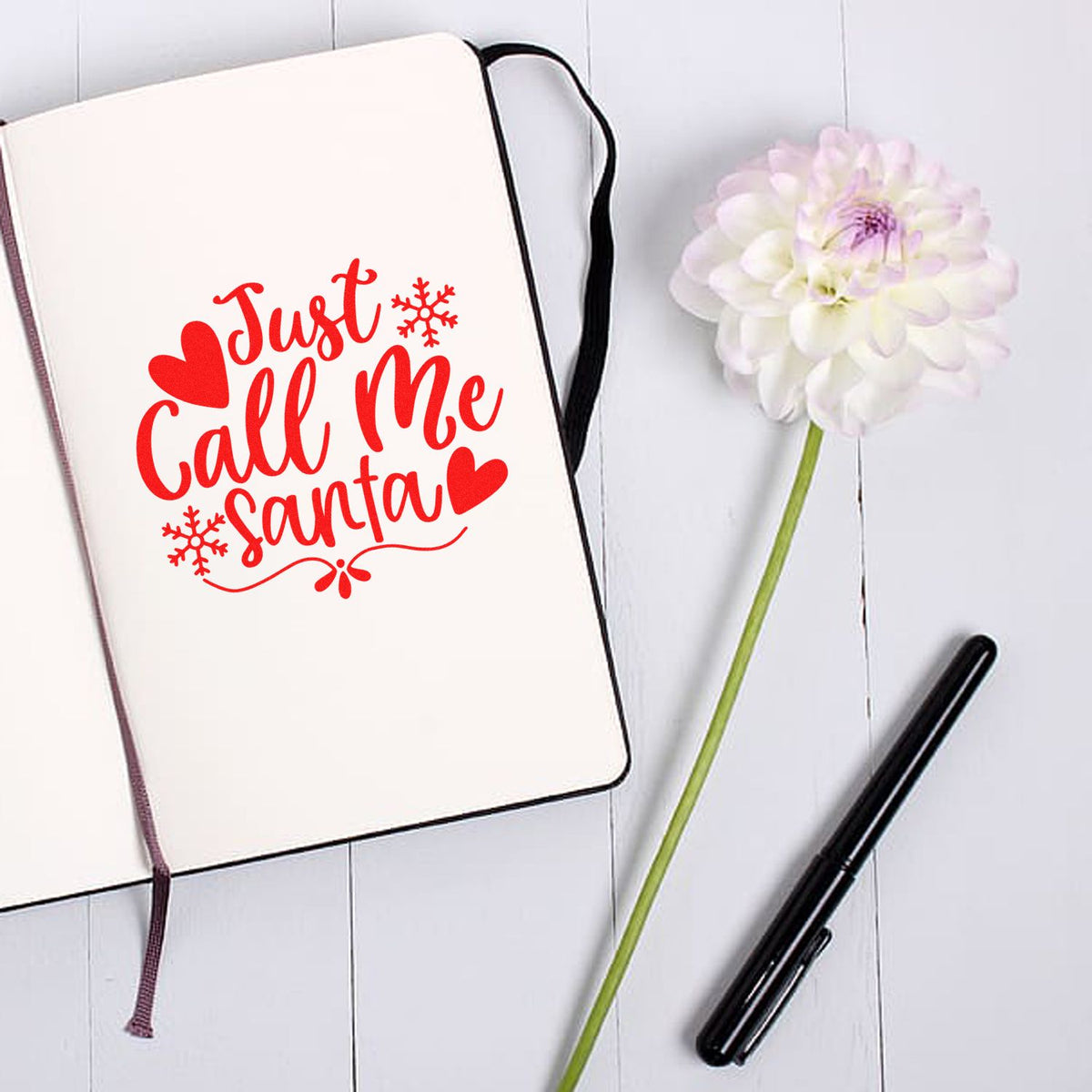 Red Just Call Me Santa stamp design on a cream notebook, with hearts, snowflakes, a white flower, and black pen.