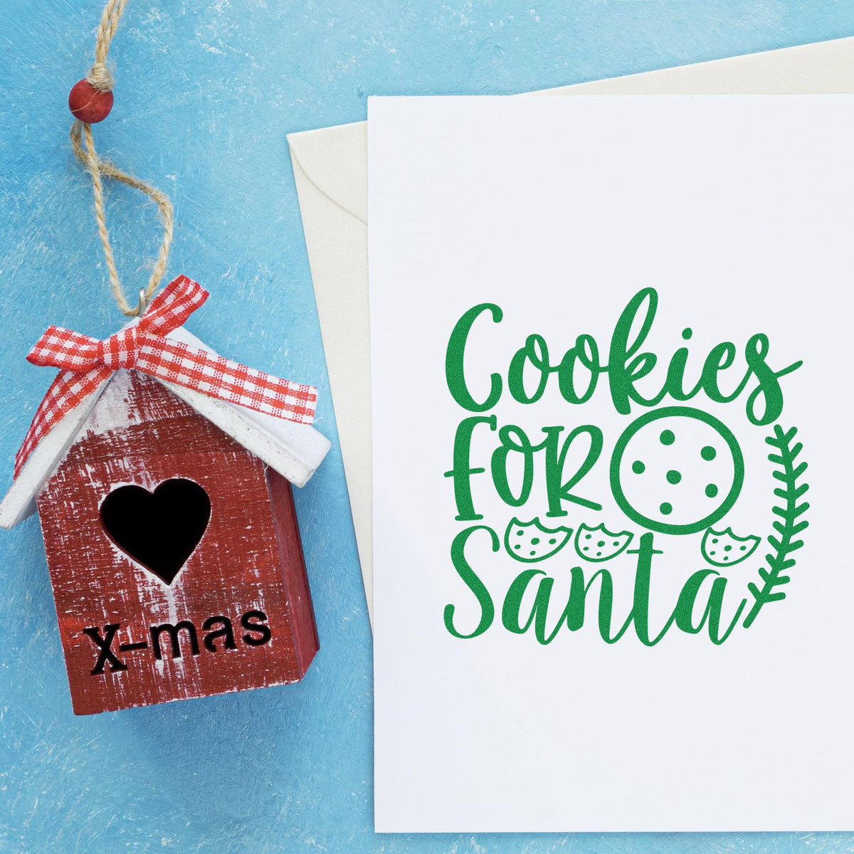 Rustic red wooden birdhouse seal ornament with heart cutout beside white card with green Cookies for Santa stamp.