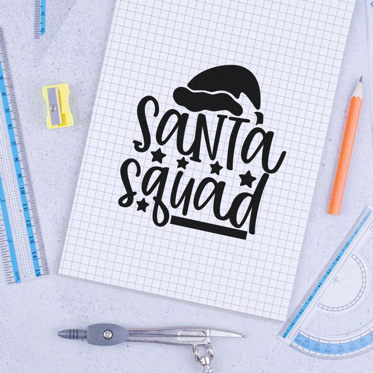 Black Santa Squad stamp design with Santa hat on graph paper, surrounded by blue rulers, pencil, and compass.