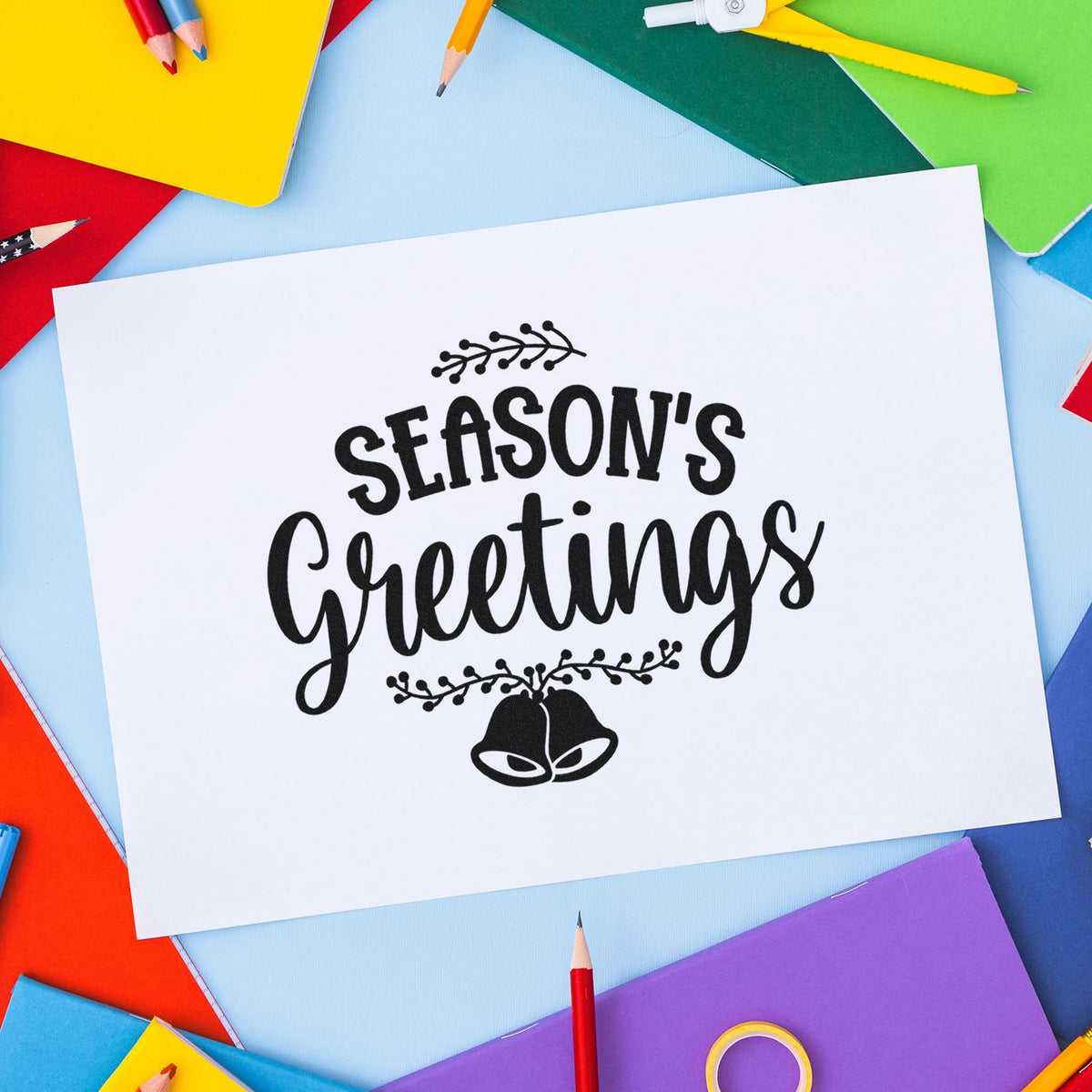 Black stamp and seal reading Seasons Greetings on white card, surrounded by bright colored notebooks and pencils.