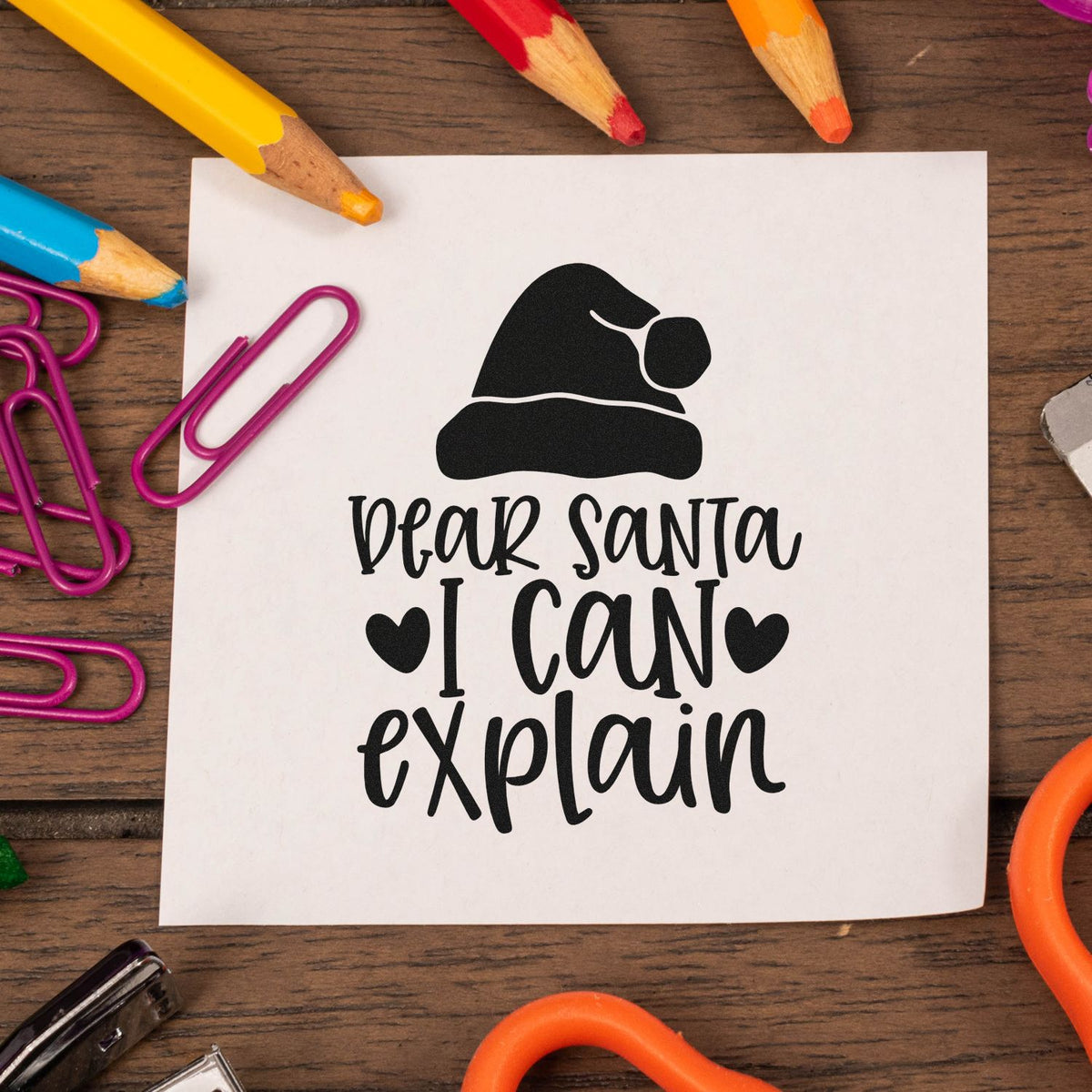Black Dear Santa I Can Explain stamp seal with Santa hat on white paper, surrounded by colorful pencils and purple paper clips.