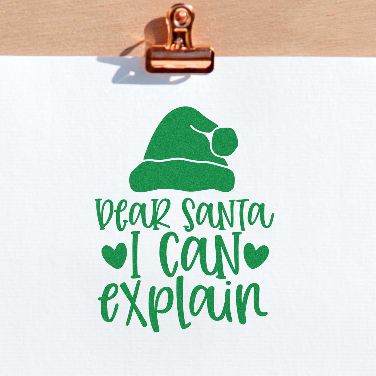Green Dear Santa I Can Explain stamp and seal design with Santa hat, hearts, on white paper clipped by copper binder clip.