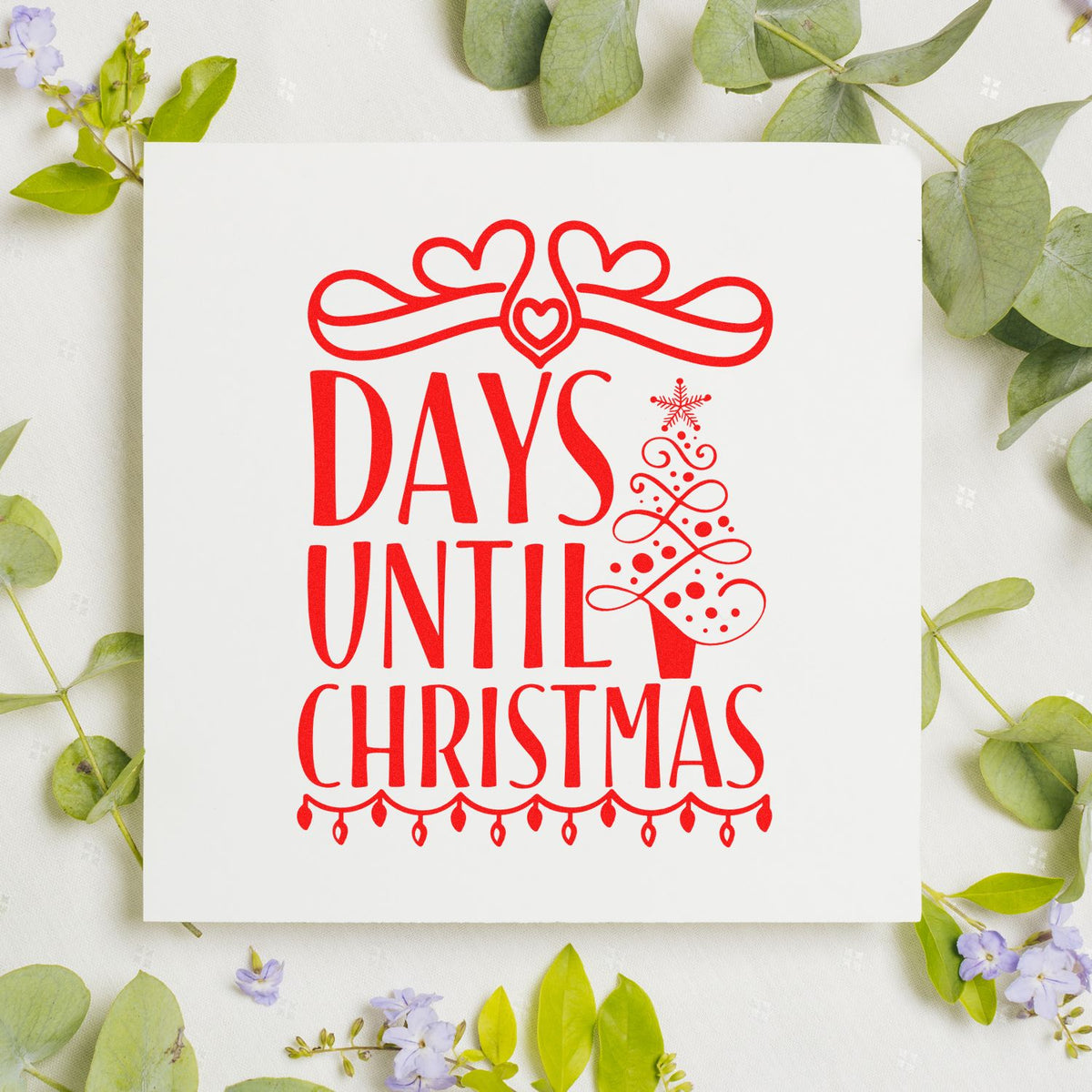Red Christmas rubber stamp seal with hearts, tree, and Days Until Christmas on white card, surrounded by green leaves.