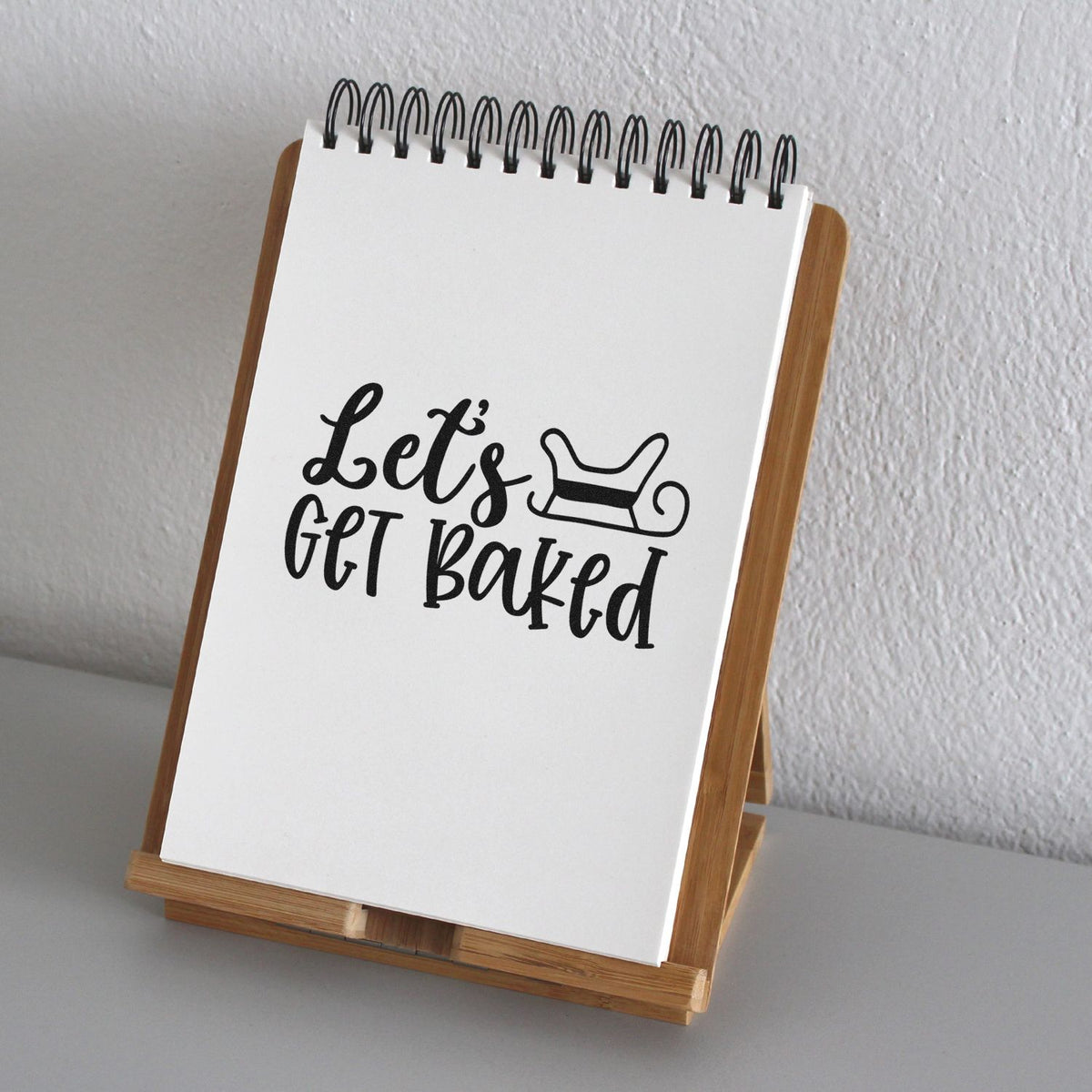 White spiral notepad on wooden stand with black Lets get baked bakery stamp and rolling pin design.