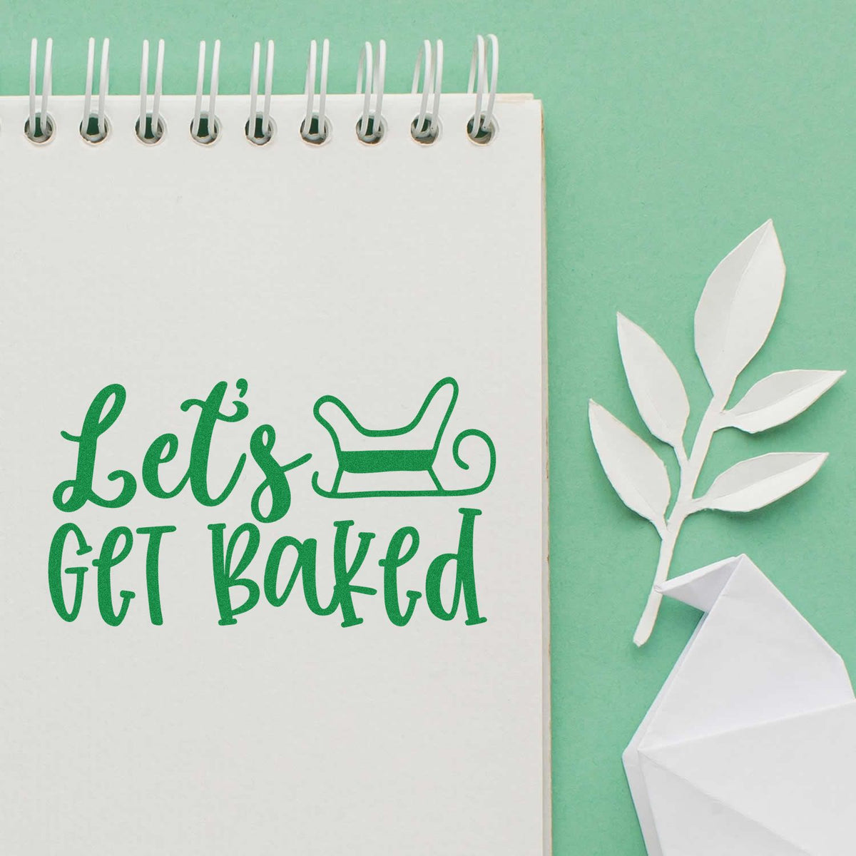 Green custom stamp and seal reading Lets Get Baked with rolling pin icon on white spiral notebook, beside white paper leaves.