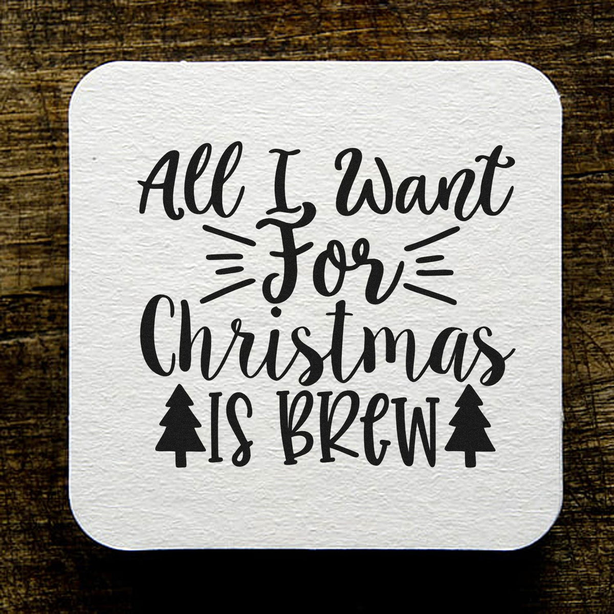 White square stamp/seal design with black All I Want For Christmas Is Brew lettering, tree icons, on rustic wood.