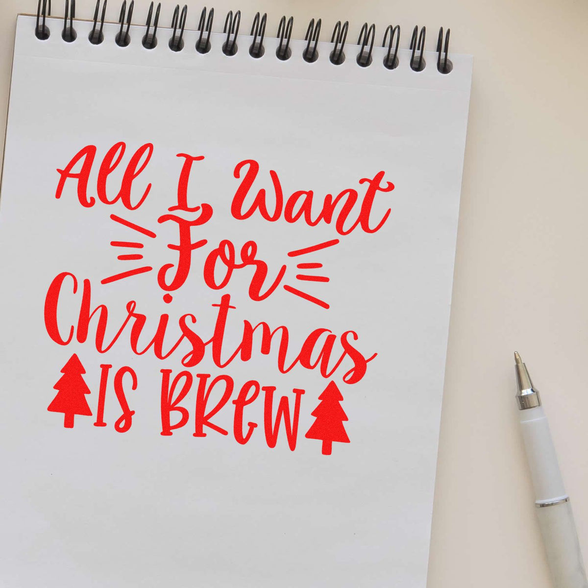Red holiday stamp reading All I Want For Christmas Is Brew on white spiral notepad, with tree icons and white pen.