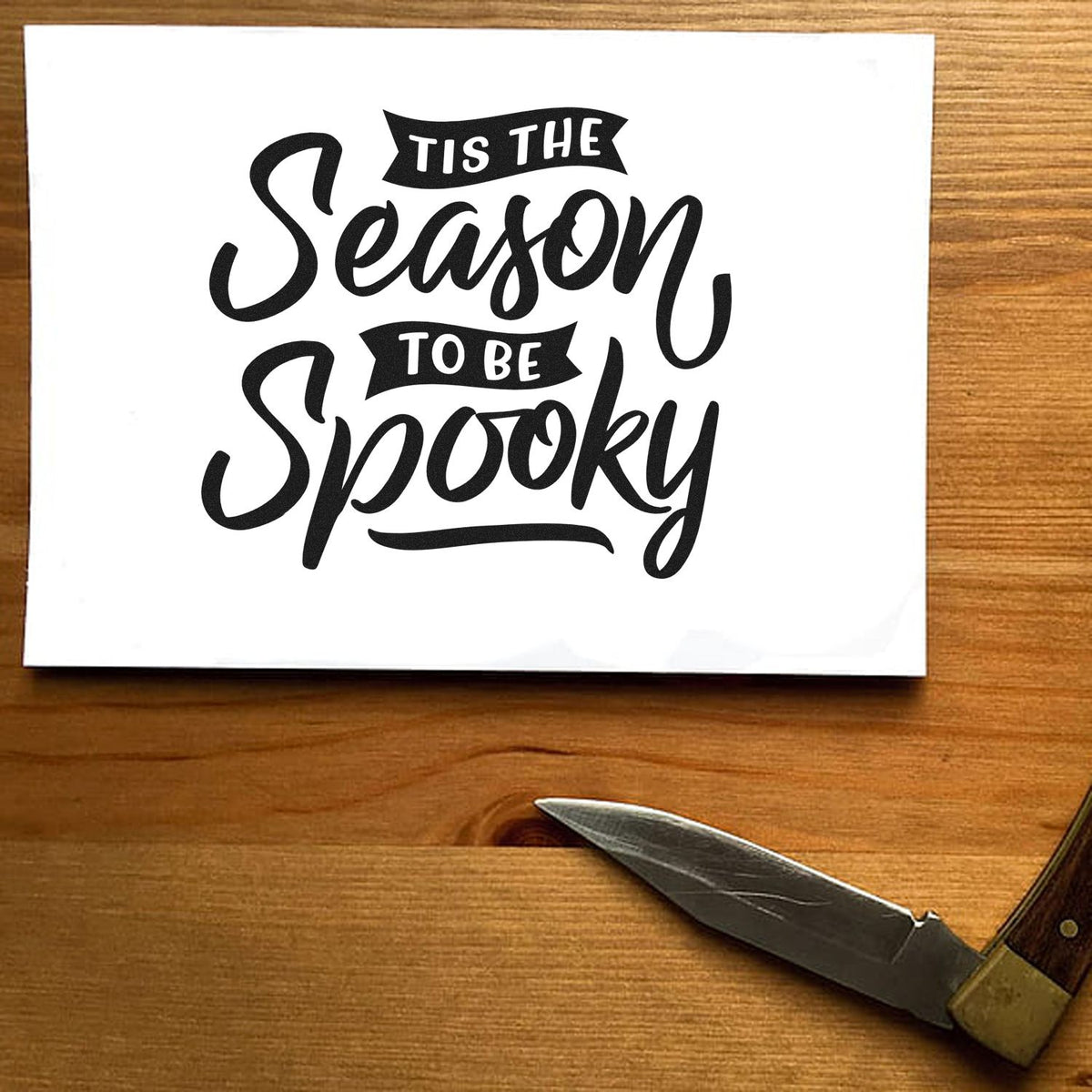 Black stamp and seal design reading Tis the Season to Be Spooky on white paper, with wood tabletop and metal tool.