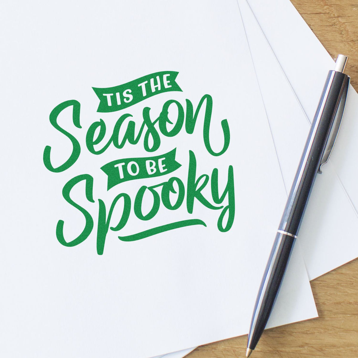 Green Tis the Season to Be Spooky stamp and seal design on white paper, with black pen on wood desk.