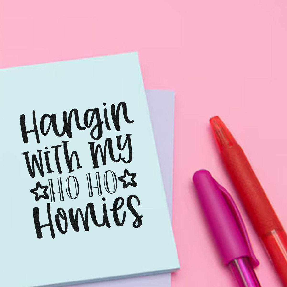 Pastel blue holiday stamp reading Hangin With My Ho Ho Homies, with stars, beside pink and red seal pens on a pink background.