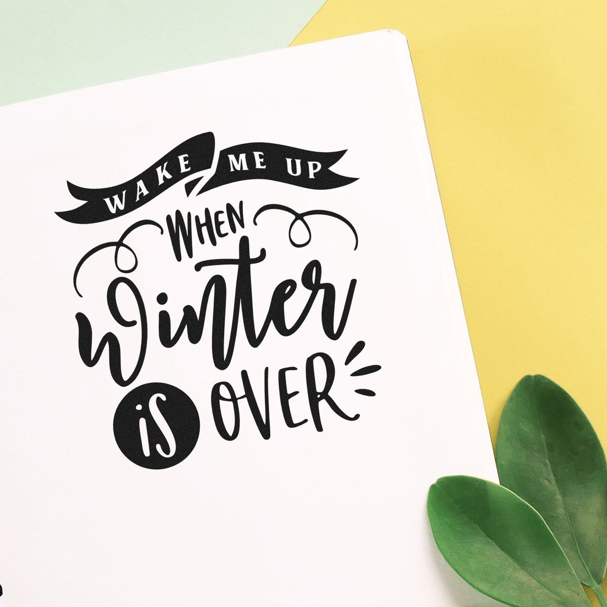 Black custom stamp design reading Wake me up when winter is over on white paper, with yellow backdrop and green leaves.