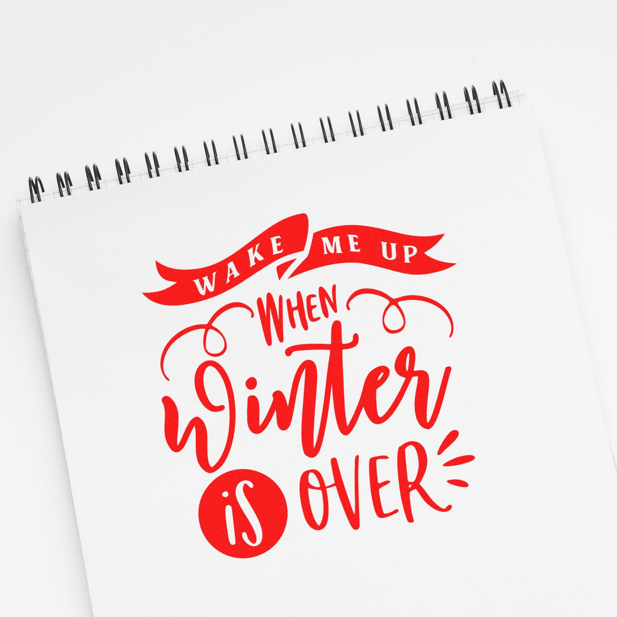 Red stamp-style lettering on a white spiral notebook reading Wake me up when winter is over.