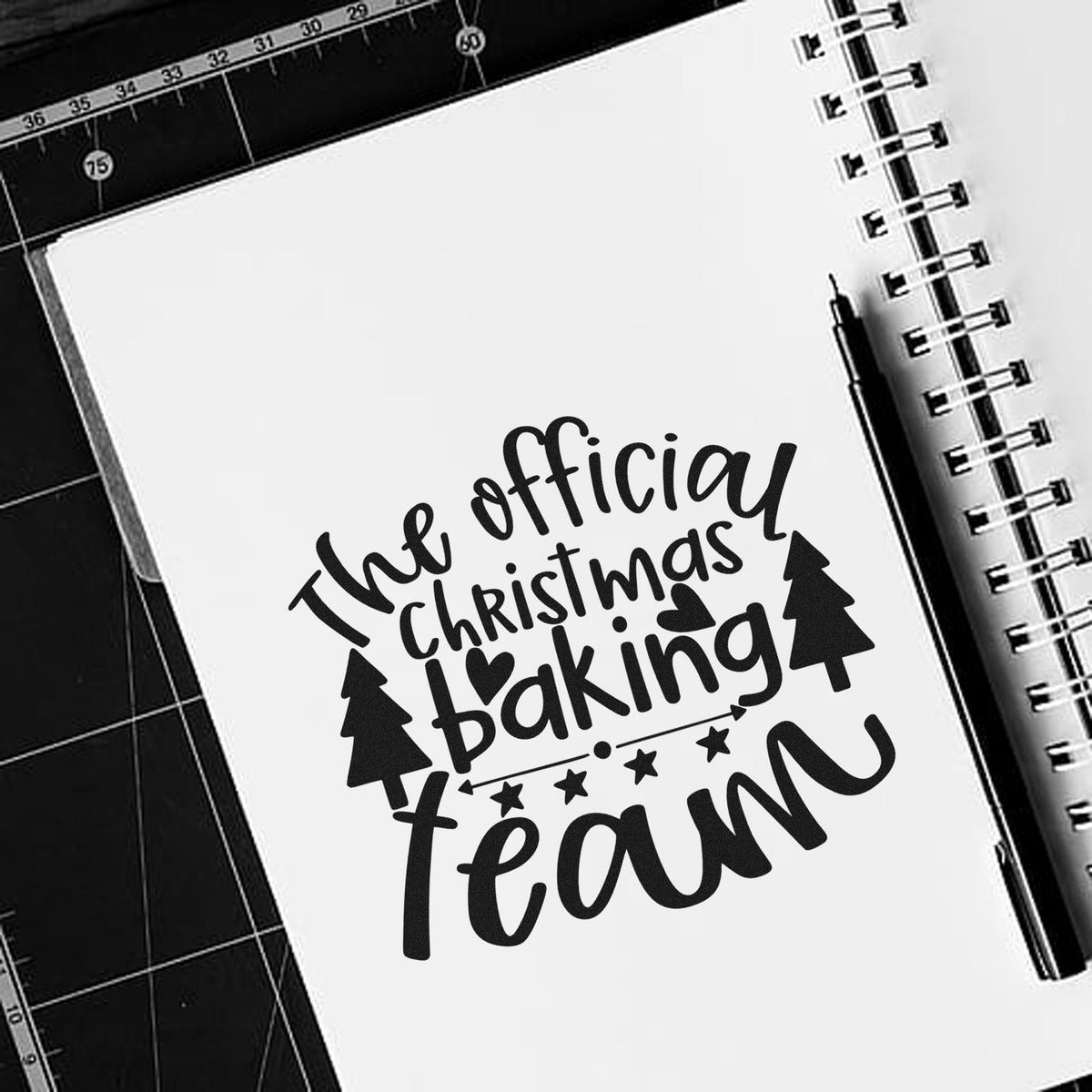Black Christmas baking team stamp and seal design on a white notebook, with trees, stars, heart, pen, and cutting mat.