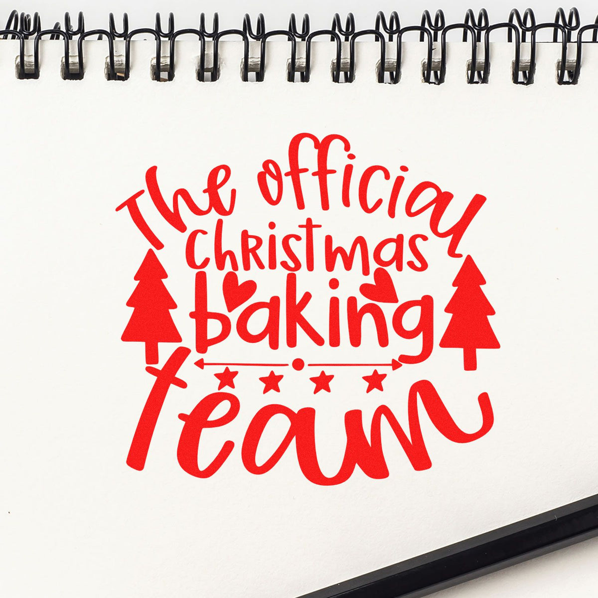Red The Official Christmas Baking Team stamp and seal design with trees, hearts and stars on a white spiral-bound page.