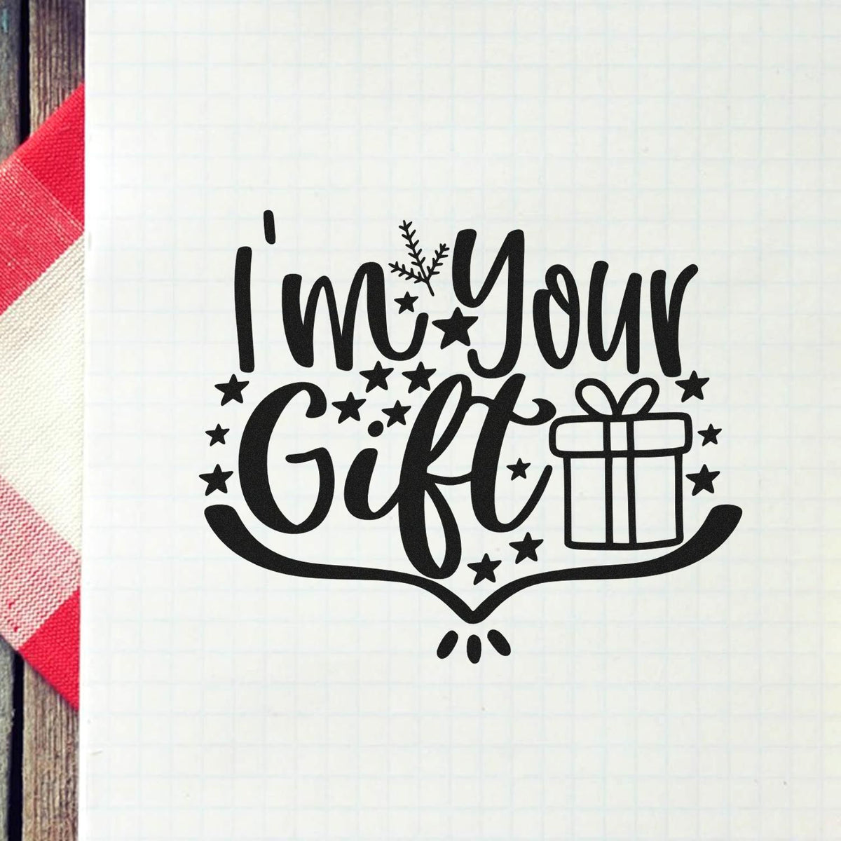 Black holiday stamp seal reading Im Your Gift with stars and a gift box on white grid paper, beside red plaid fabric.