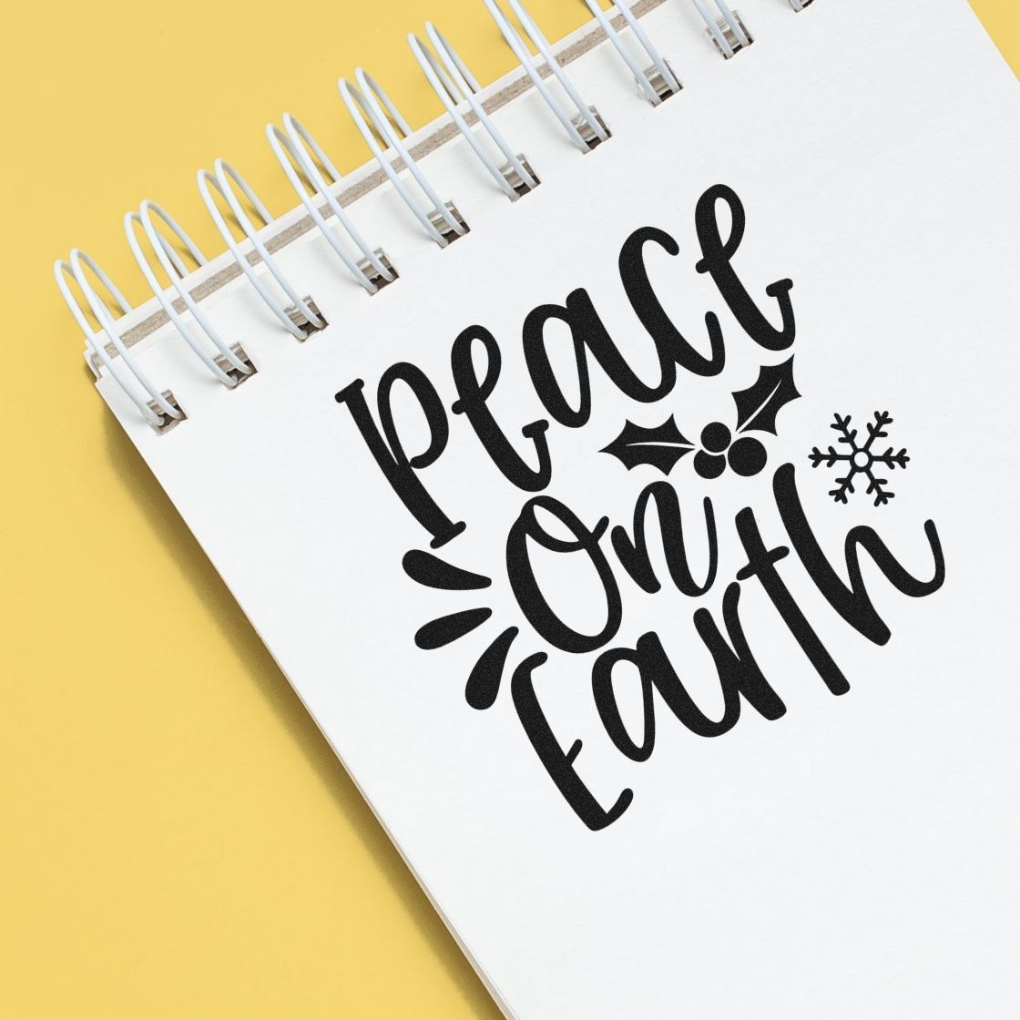 Black peace on earth holiday stamp and seal design with holly and snowflake on a white spiral notebook, yellow background.