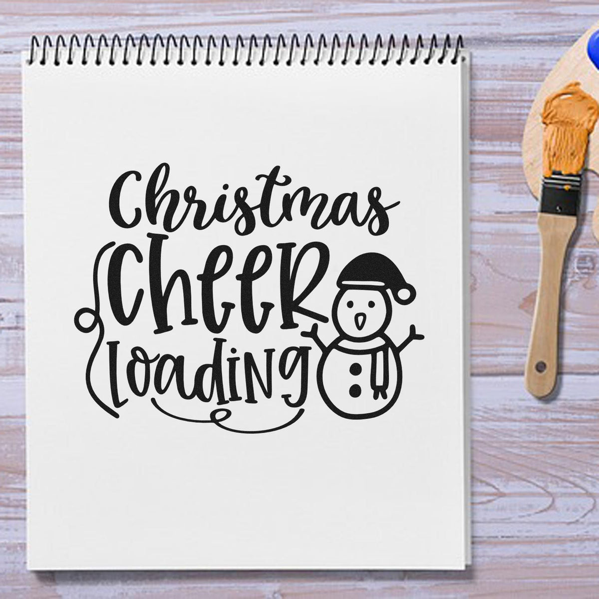 White spiral notebook with black Christmas Cheer Loading stamp design, snowman, and wooden seal stamp with orange ink.