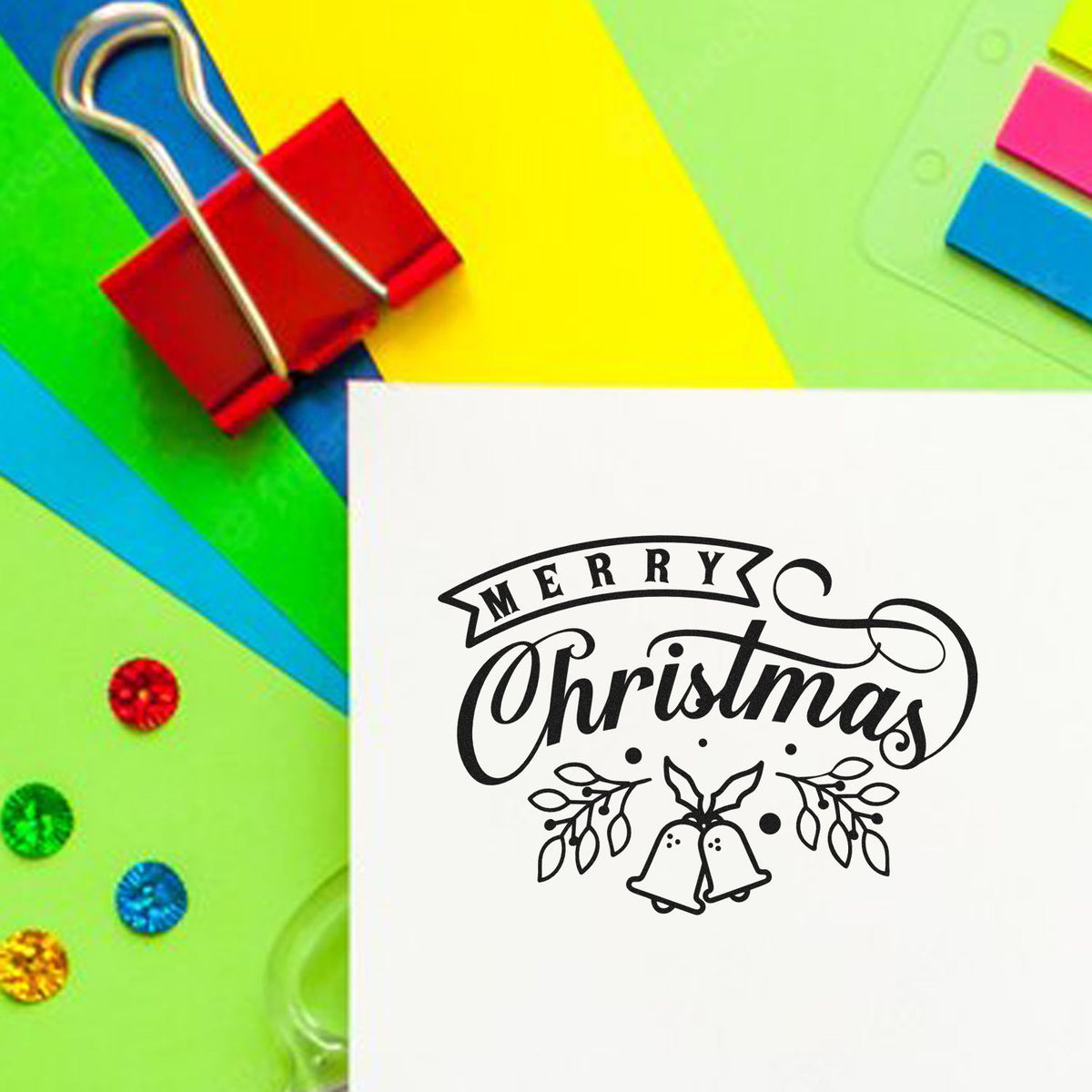 Merry Christmas stamp and seal design with bells on white paper, on bright green, yellow, blue, and red craft background.