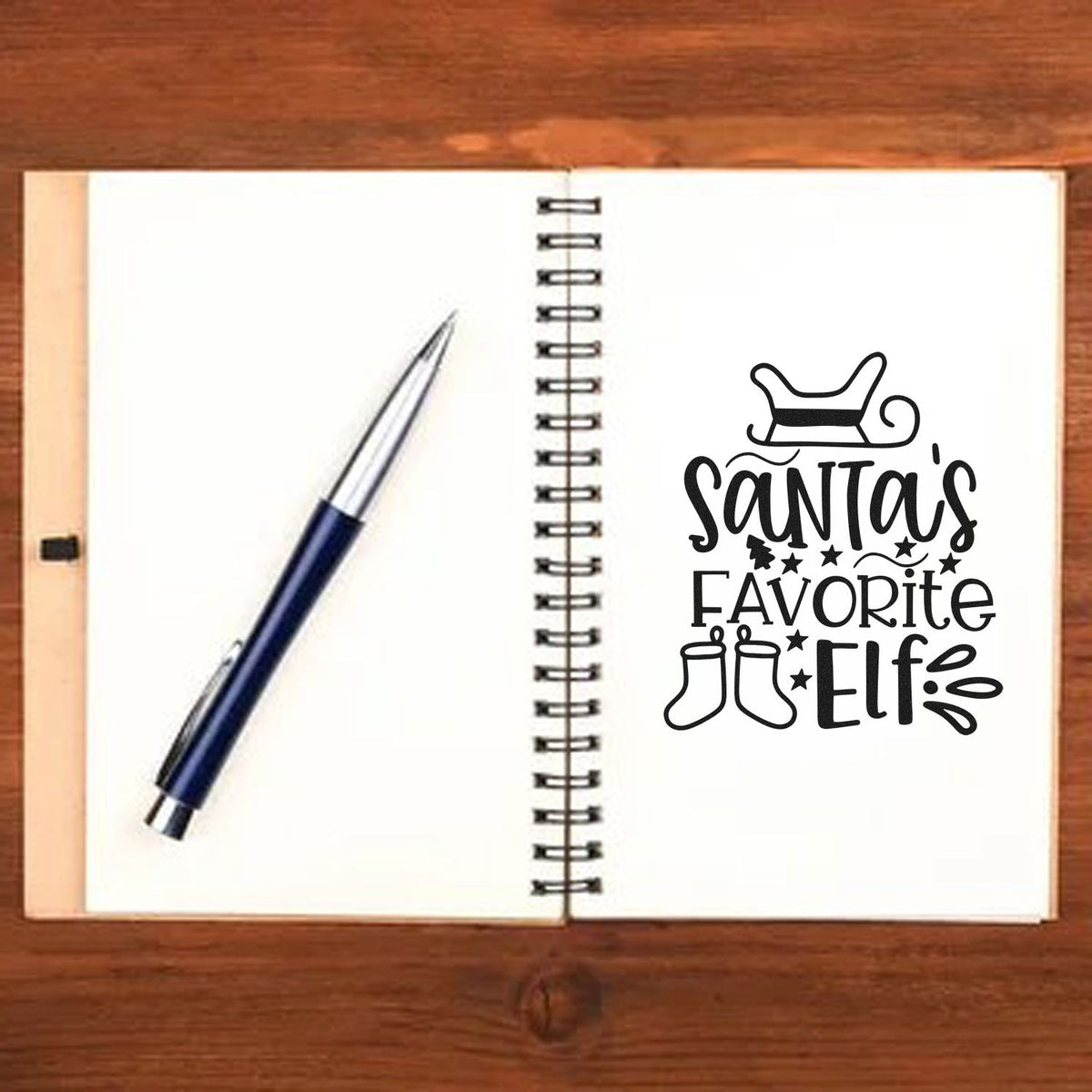 Open notebook showing a black Santas Favorite Elf stamp seal design with hat, stars, boots, and a blue pen.