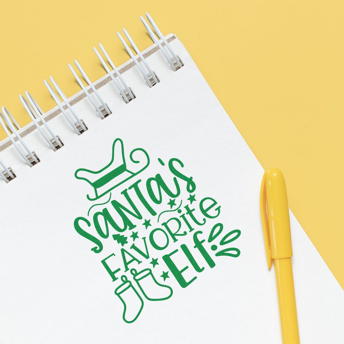 Green Santas Favorite Elf! stamp design on white spiral notebook with yellow pen and pastel yellow background.