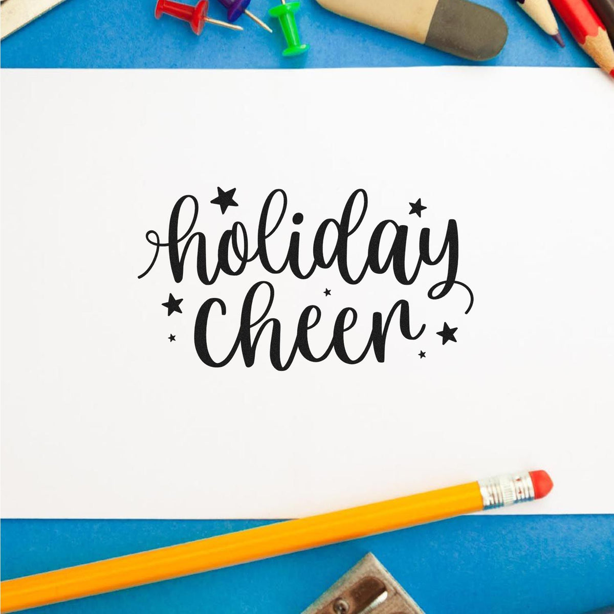 Black holiday cheer stamp design on white card, framed by colorful pencils, push pins, and blue background.
