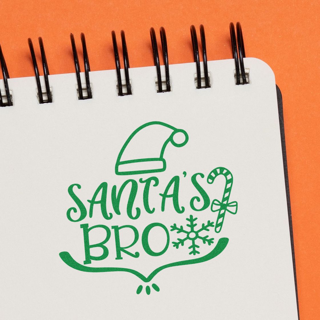 Green Santas Bro stamp and seal design with Santa hat, candy cane and snowflake on a white notebook page, orange background.
