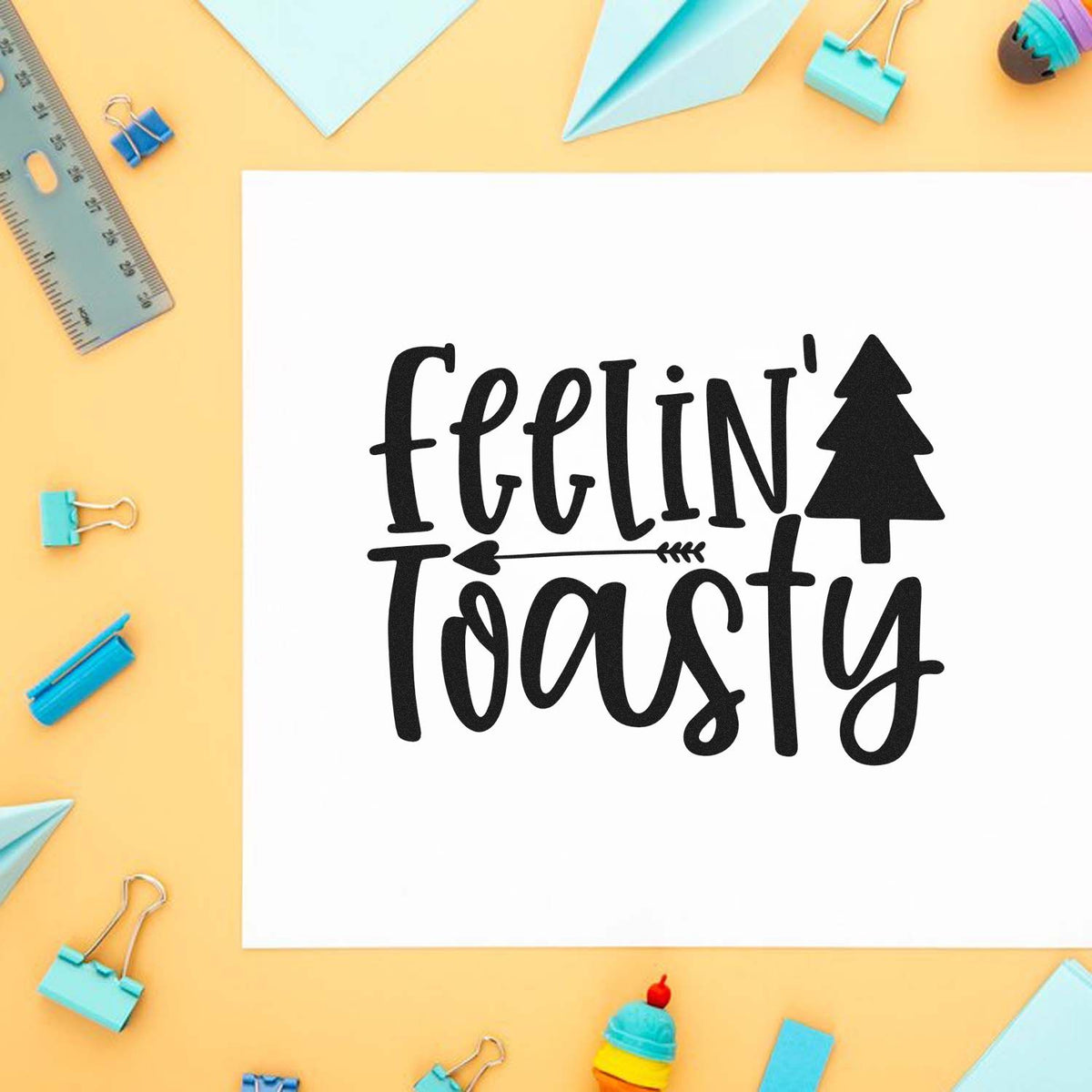Black 'feelin' toasty' stamp on white paper with pine tree, on peach background with pastel blue clips, ruler, and paper planes.