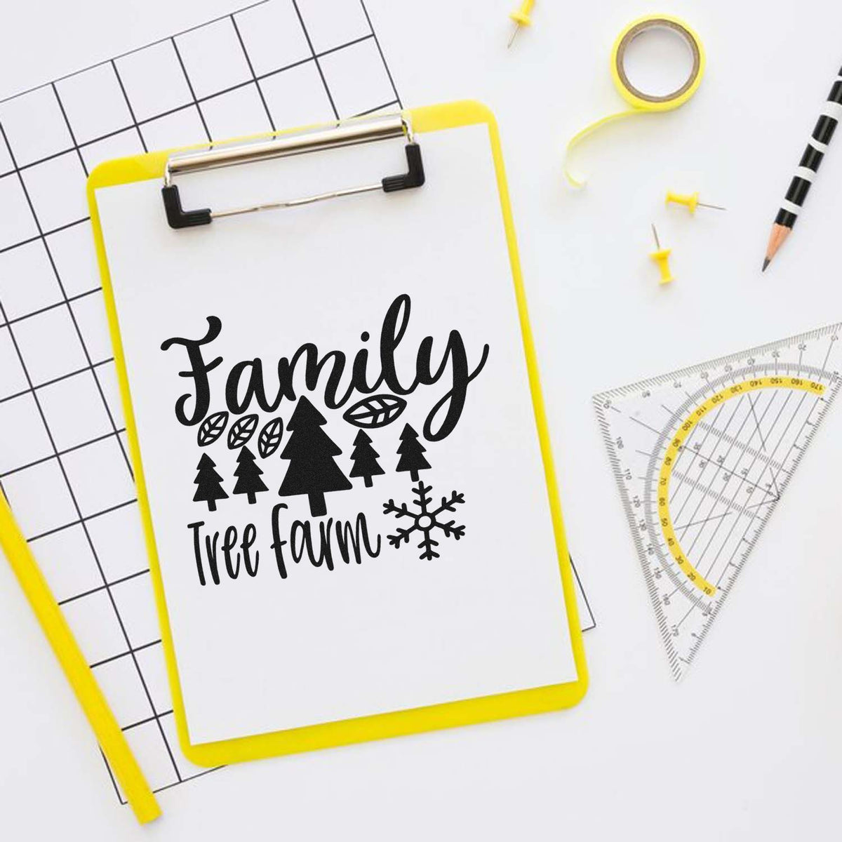 Yellow clipboard displaying black Family Tree Farm stamp with trees and snowflake, beside yellow tape, pins, and a ruler.