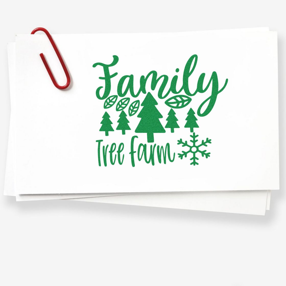 Green Family Tree Farm stamp design in green on white paper, with pine trees and snowflake, clipped by a red paper clip.